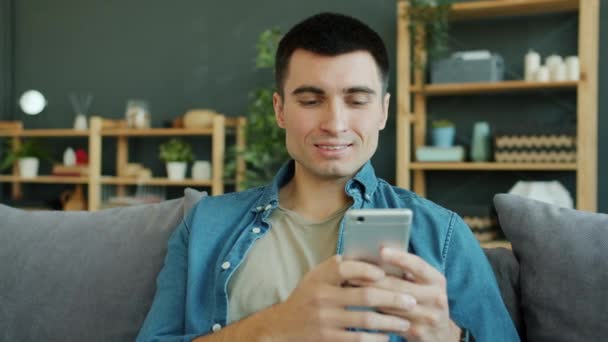 Joyful man using smartphone texting having fun smiling in house alone - Filmati, video