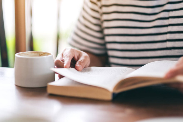 Крупный план женщины, держащей и читающей книгу с кофейной чашкой на деревянном столе - Фото, изображение