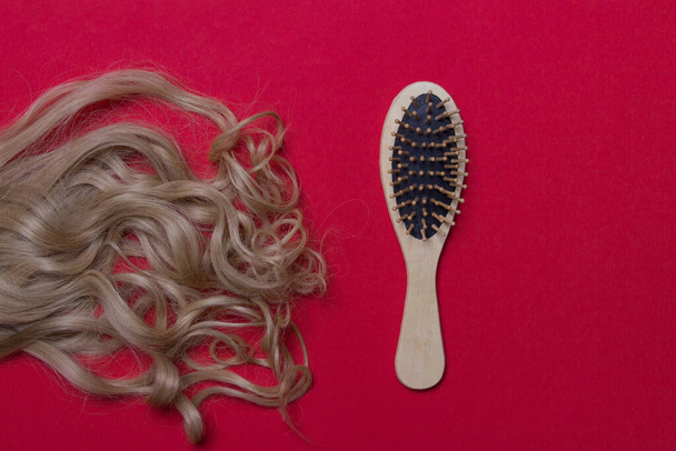 lock of woman blond hair and wooden hairbrush on red colored paper background. - Photo, Image