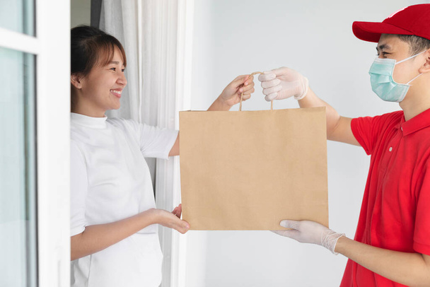 Azji dostawca trzyma torbę na zakupy w medycznych rękawic gumowych i maski dać młodej kobiecie przed domem, - Zdjęcie, obraz
