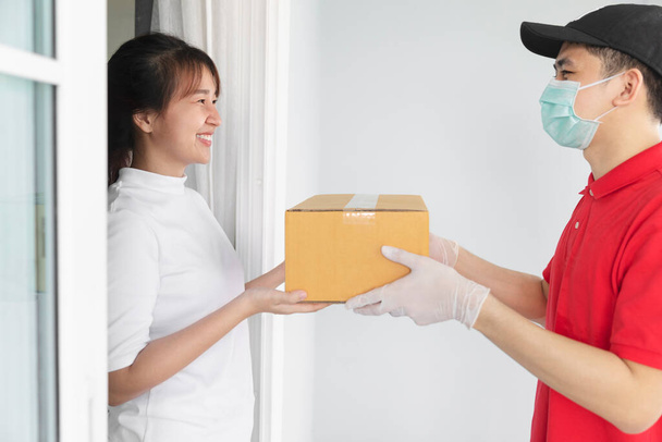 Azji dostawca trzyma kartonowe pudełka w medycznych rękawic gumowych i maski dać młodej kobiecie przed domem, - Zdjęcie, obraz