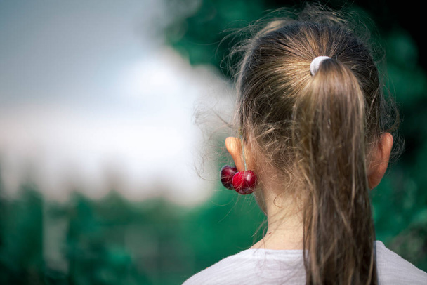 Beautiful little girl got a fresh cherry on her ear. Adorable little girl with a cherry on her ear. Making jewelry from fruit. - Fotografie, Obrázek