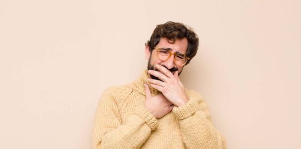 young cool man feeling ill with a sore throat and flu symptoms, coughing with mouth covered against flat wall - Photo, Image