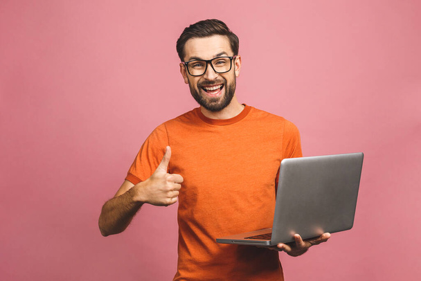 Selbstbewusster Wirtschaftsexperte. Selbstbewusster junger gutaussehender Mann in lässiger Haltung, Laptop in der Hand und lächelnd vor rosafarbenem Hintergrund. Daumen hoch. - Foto, Bild