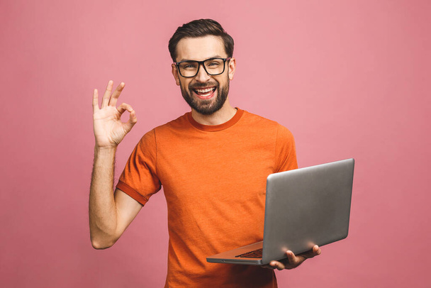 Zelfverzekerde zakenexpert. Zelfverzekerde jonge knappe man in casual vasthouden laptop en glimlachen terwijl staan tegen roze achtergrond. Ok teken. - Foto, afbeelding