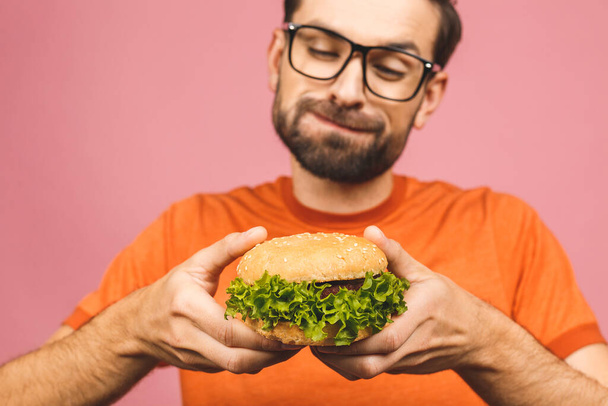 Young man holding a piece of hamburger. Student eats fast food. Burger is not helpful food. Very hungry guy. Diet concept. Isolated over pink background. - Fotografie, Obrázek