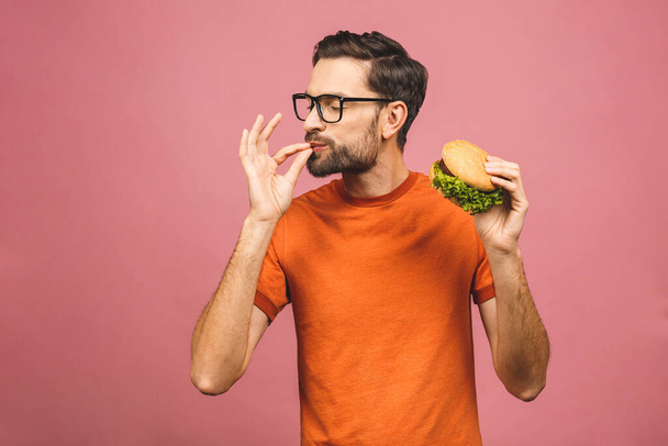 Young man holding a piece of hamburger. Student eats fast food. Burger is not helpful food. Very hungry guy. Diet concept. Isolated over pink background. - Photo, Image