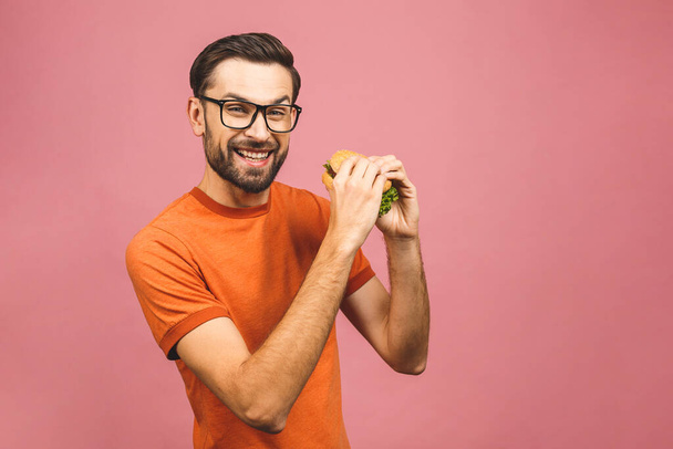 Young man holding a piece of hamburger. Student eats fast food. Burger is not helpful food. Very hungry guy. Diet concept. Isolated over pink background. - Photo, Image