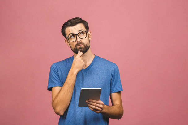Thinking serious young man in casual standing and using tablet over pink background - Fotó, kép