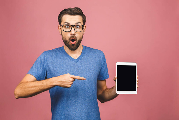 Barbuto uomo stupito in occhiali da vista che mostrano schermo del computer tablet vuoto e guardando la fotocamera. isolato su sfondo rosa
.  - Foto, immagini
