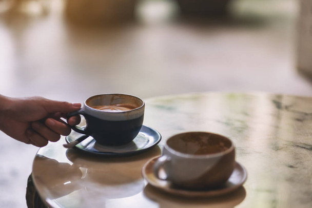 Zbliżenie obraz dłoni trzymającej niebieską filiżankę gorącej kawy z innym kubkiem na marmurowym stole w kawiarni - Zdjęcie, obraz