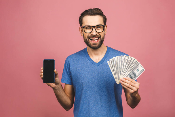 Ενθουσιασμένος άνθρωπος σε casual t-shirt κρατώντας πολλά χρήματα σε δολάρια νομίσματα και το κινητό τηλέφωνο σε χέρια που απομονώνονται πάνω από ροζ τοίχο. Τηλεφωνική οθόνη για κείμενο.  - Φωτογραφία, εικόνα