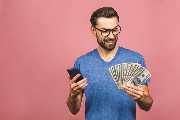Ενθουσιασμένος άνθρωπος σε casual t-shirt κρατώντας πολλά χρήματα σε δολάρια νομίσματα και το κινητό τηλέφωνο σε χέρια που απομονώνονται πάνω από ροζ τοίχο. - Φωτογραφία, εικόνα