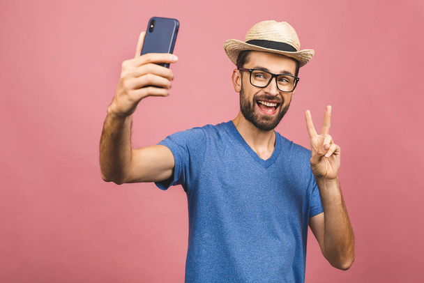 Концепция путешествия. Портрет веселого молодого человека в соломенной шляпе, делающего селфи со смартфоном на розовом фоне
.  - Фото, изображение