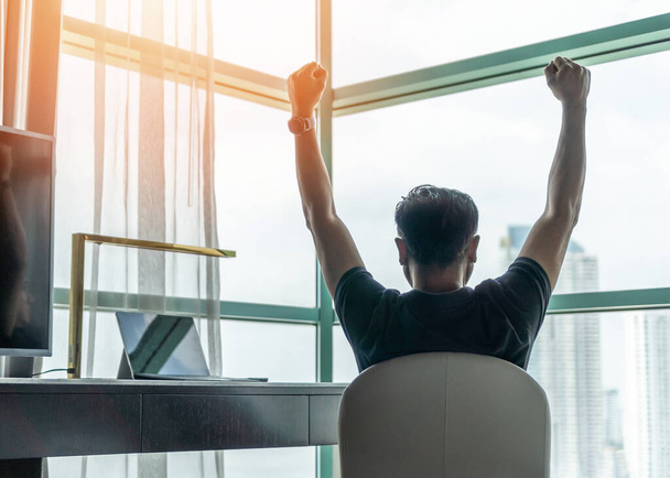 Business achievement concept with happy businessman relaxing in office or hotel room, resting and raising fists with ambition looking forward to city building urban scene through glass window - Photo, Image