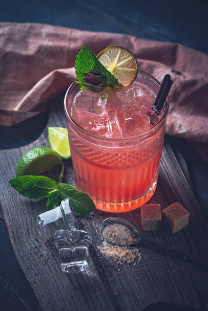 Бокал красного алкогольного коктейля со льдом, мятой и лаймом. Летний освежающий напиток на темном фоне
 - Фото, изображение