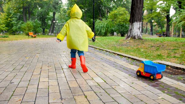 Χαριτωμένο αγοράκι με κίτρινο αδιάβροχο τρέχει με φορτηγό παιχνίδι κάτω από τη βροχή στο πάρκο - Φωτογραφία, εικόνα