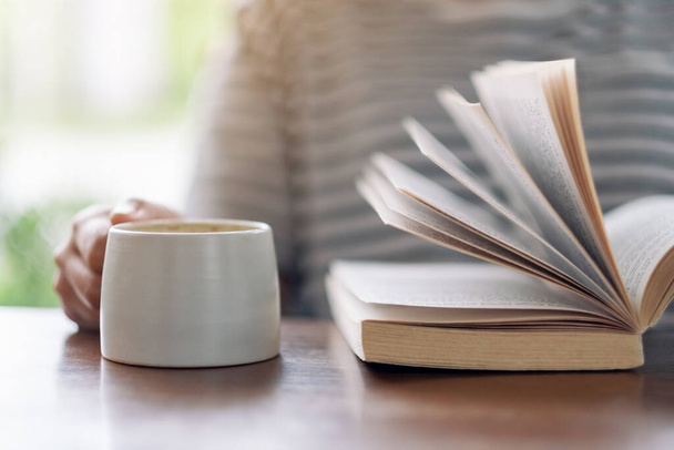 Image rapprochée d'une femme tenant et lisant un livre tout en buvant du café sur une table en bois - Photo, image