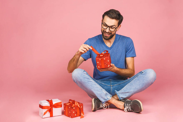 Bezauberndes Foto von attraktiven jungen Mann mit schönem Lächeln hält Geburtstagsgeschenkboxen isoliert über rosa Hintergrund, auf dem Boden sitzend. Geschenkkonzept.  - Foto, Bild