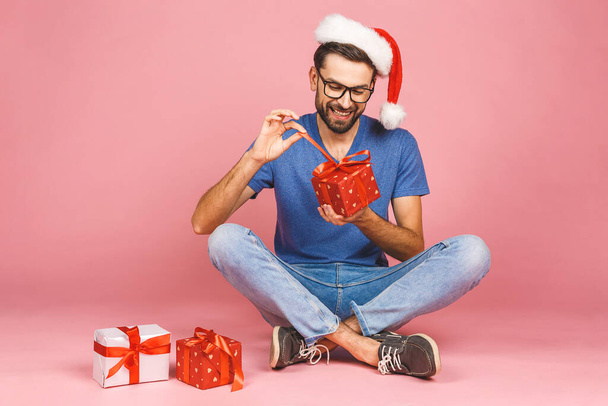 Adorable photo of attractive young man in christmas hat with beautiful smile holding birthday present boxes isolated over pink background, sitting on the floor. Gift concept. - Photo, Image