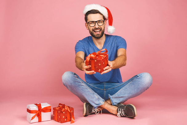 ピンクの背景に隔離された美しい笑顔を保持誕生日プレゼントボックスとクリスマスの帽子の魅力的な若い男の愛らしい写真は、床に座っています。贈り物のコンセプト. - 写真・画像