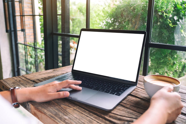Mockup-Bild einer Hand, die beim Kaffeetrinken ein Laptop-Touchpad mit leerem weißen Desktop-Bildschirm benutzt und berührt - Foto, Bild