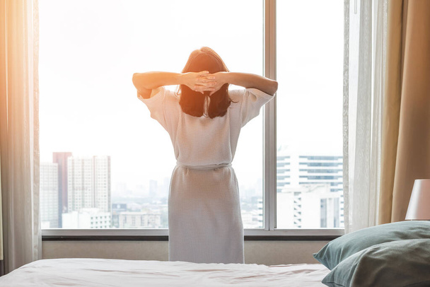 Helppo elämäntapa Aasialainen nainen herää hyvästä unesta viikonloppuna aamulla ottaen lepoa, rentoutuen mukavassa makuuhuoneessa hotellin ikkunassa, nauttien onnellisesta laiskasta päivästä nauttien työn ja elämän laadun tasapainosta. - Valokuva, kuva