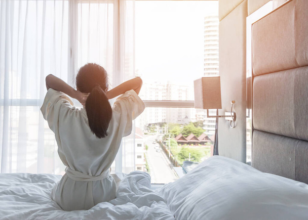 Easy Lifestyle Asiatische Frau, die am Wochenende morgens aus dem Schlaf erwacht und sich ausgeruht hat, entspannt sich im komfortablen Schlafzimmer am Hotelfenster, genießt einen fröhlichen faulen Tag und genießt das Work-Life-Balance-Konzept - Foto, Bild