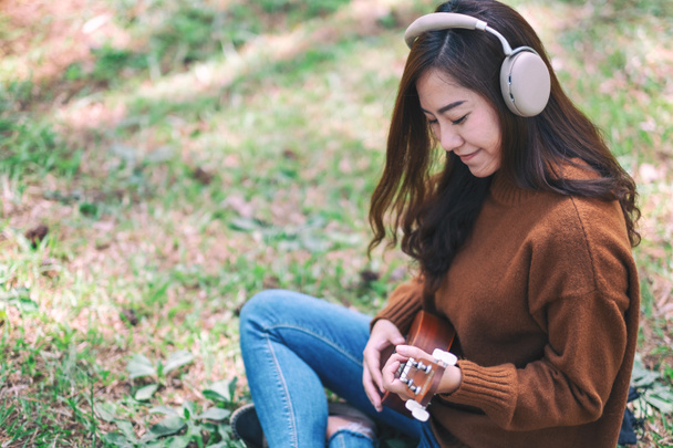 Una hermosa mujer asiática disfruta escuchando música con auriculares mientras juega ukelele en el parque
 - Foto, Imagen