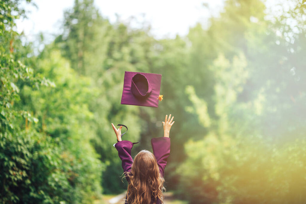 Foto von hinten Absolvent Abschluss kleines Mädchen mit Mütze werfen es in den Himmel, glücklich über das Ende, violette Kostüm. - Foto, Bild