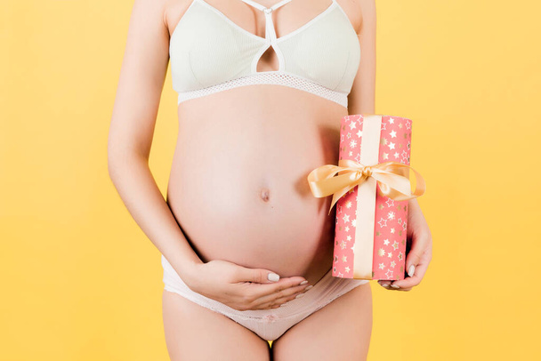 Cropped image of pregnant woman in white underwear holding a gift box and touching her belly at blue background. Expecting a baby girl. Copy space - Fotografie, Obrázek