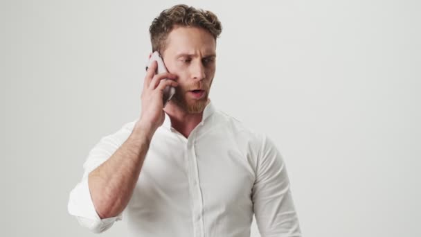 Handsome young concentrated man in white shirt isolated over white wall background talking by mobile phone - Imágenes, Vídeo