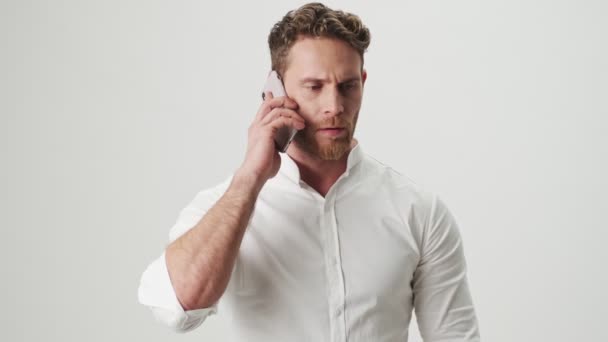 Handsome young angry screaming man in white shirt isolated over white wall background talking by mobile phone - Imágenes, Vídeo