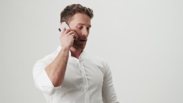 Handsome young optimistic man in white shirt isolated over white wall background talking by mobile phone make winner gesture - Séquence, vidéo