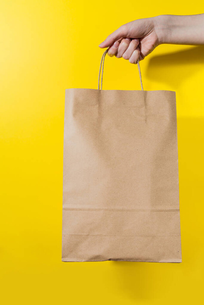 mano femenina sostiene una bolsa de papel con mercancías sobre un fondo amarillo. comprar bienes en línea. entrega de mercancías. Cartel publicitario
. - Foto, imagen