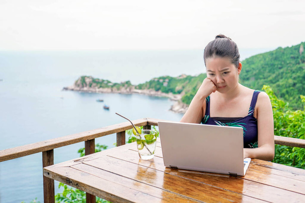 Азиатская девушка напряженно работает в режиме онлайн на фоне вида на море с балкона над уровнем моря
 - Фото, изображение