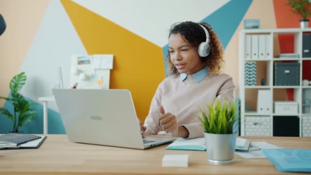 Afro-American lady in headphones listening to music and using laptop in modern office - Záběry, video
