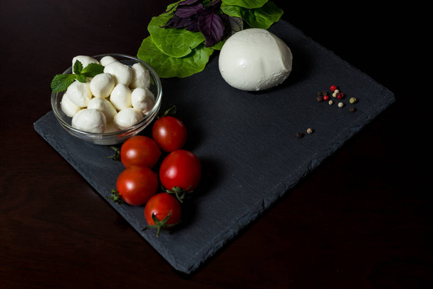 Lebensmittel-Hintergrund. Große und kleine Mozzarella-Bällchen auf einem Steinteller mit Basilikumblättern, Salat, Tomaten und Paprika. Auf dem alten Hintergrund. Freier Kopierraum. Ansicht von oben. Konzept für Menügestaltung - Foto, Bild