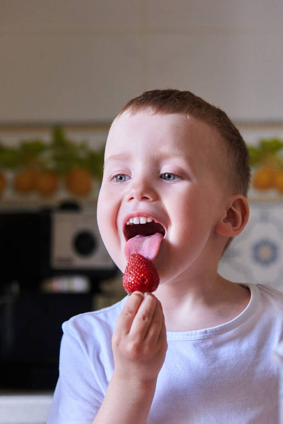 Beautiful European child in the kitchen eating strawberries and drinking milk. - Fotografie, Obrázek