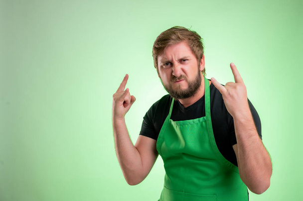 Yeşil önlüklü ve siyah tişörtlü bir süpermarket çalışanı yeşil arka planda elleri yukarıda rock sembolü yapıyor. - Fotoğraf, Görsel