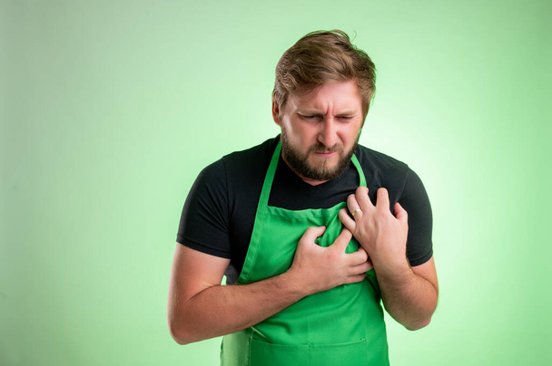 Supermarket employee with green apron and black t-shirt, suffering heart ache or breast pain, cardiac problems isolated on green background - Photo, Image
