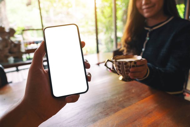 Mockup εικόνα του χεριού ενός άνδρα κρατώντας μαύρο κινητό τηλέφωνο με λευκή οθόνη με γυναίκα πίνοντας καφέ στο καφέ - Φωτογραφία, εικόνα