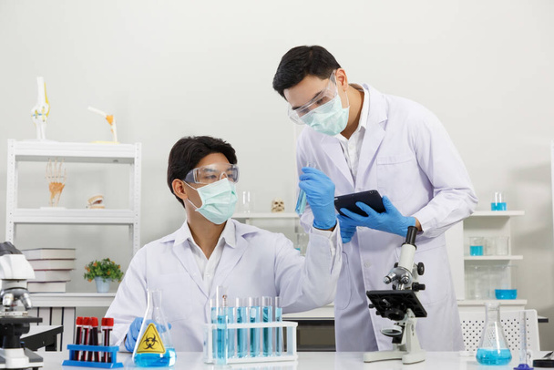 Δύο αρσενικοί επιστήμονες που εργάζονται στο εργαστήριο ενώ ελέγχουν το αποτέλεσμα των δοκιμασιών δειγμάτων. Μπλε τόνος. SARS-CoV-2, Covid-19 ΘΕΜΑ. - Φωτογραφία, εικόνα