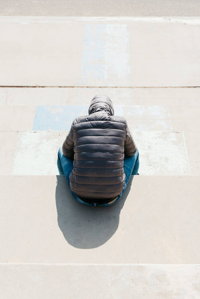 Kot pantolon ve gri kapüşonlu ceket giyen beyaz bir adam, yukarıdan görülmüş, beton bir merdivende oturuyor. - Fotoğraf, Görsel