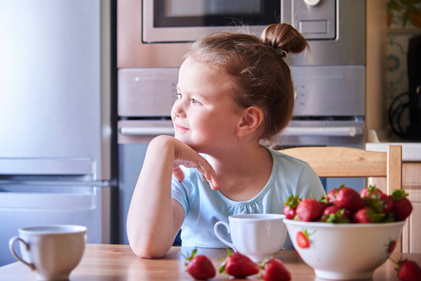 Beautiful European child in the kitchen eating strawberries and drinking milk. - Zdjęcie, obraz
