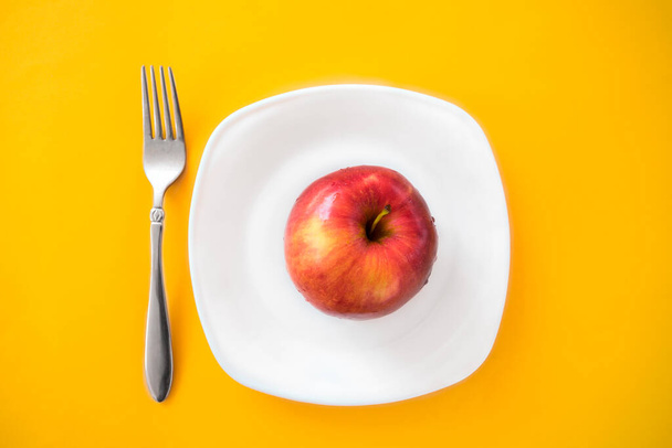 red ripe apple lies in a white plate with a fork on a yellow background. Proper nutrition. Diet. the right way of life. minimalist photo about food. - Photo, Image