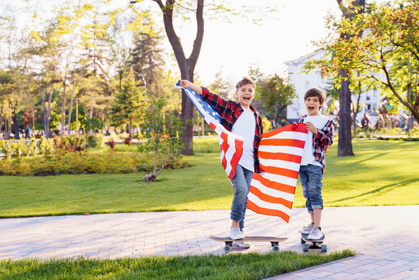 America's USA Independence Day. Two cute brothers guy americans standing in a city park on skateboards and holding flag of USA. Stretching fabric and shouting exiting with joy in flannel shirts. - Photo, Image