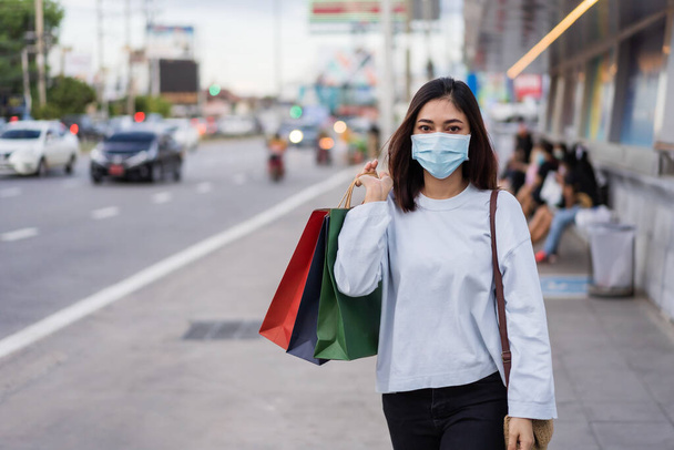 young woman holding shopping bag at bus stop in city street and wearing face mask protective for spreading of coronavirus(covid-19) pandemic, new normal concept - Fotografie, Obrázek