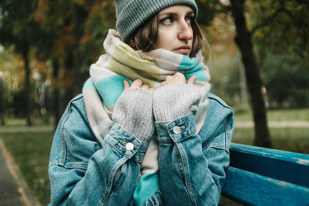 Lifestyle autumn portrait of young adult woman sitting on a bench wrapped herself in warm scarf - Foto, Bild