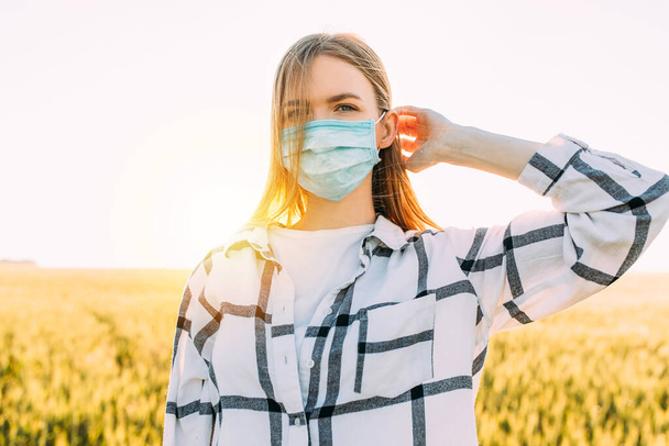 beautiful woman in a checked shirt and a protective medical mask on her face stands in a wheat field at sunset. Quarantine, coronavirus, crop - Foto, immagini